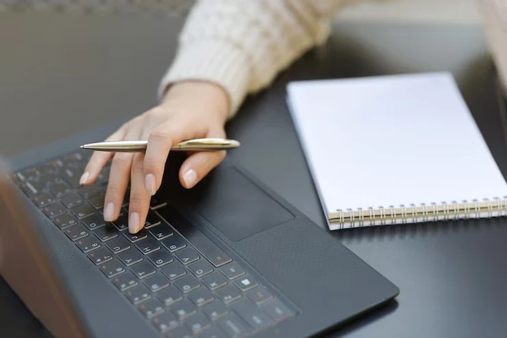 Pessoa escrevendo em um laptop enquanto segura uma caneta, com um bloco de notas ao lado, representando a criação de conteúdo longo e curto.