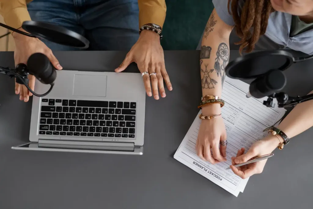 Duas pessoas trabalhando juntas na evolução do jornalismo de dados, analisando informações em uma mesa com laptop, microfones e uma planilha. Importância na era da desinformação
